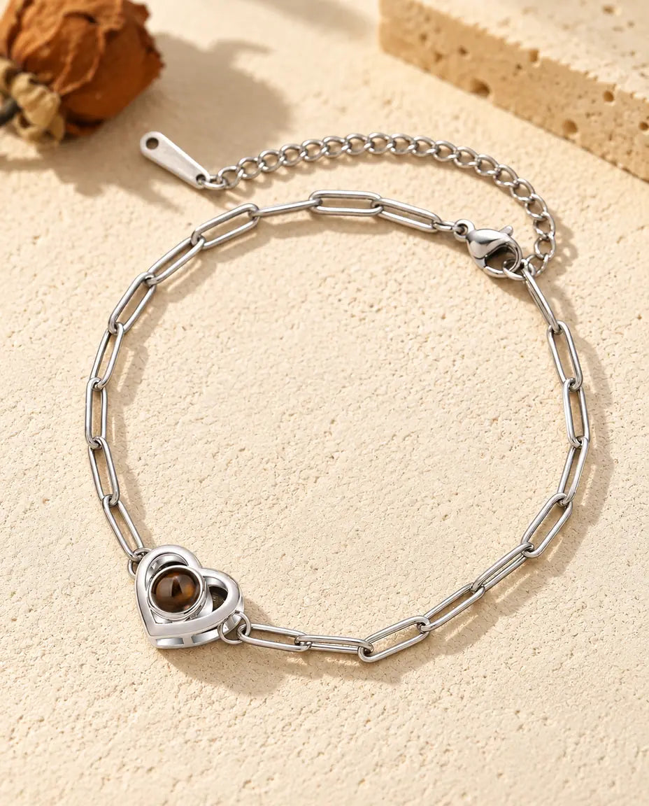 Zilverkleurige armband met foto erin en een dubbele hartbedel aan een paperclipketting, op crèmekleurig gips