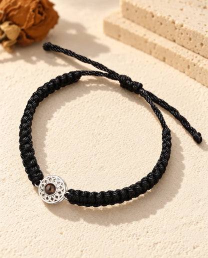 Armband met foto erin: opengewerkte zilverkleurige ronde bedel aan een zwart, breed gevlochten koord, op crèmekleurig gips