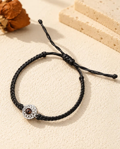 Armband met foto erin: opengewerkte zilverkleurige ronde bedel aan een zwart, dun gevlochten koord, op crèmekleurig gips