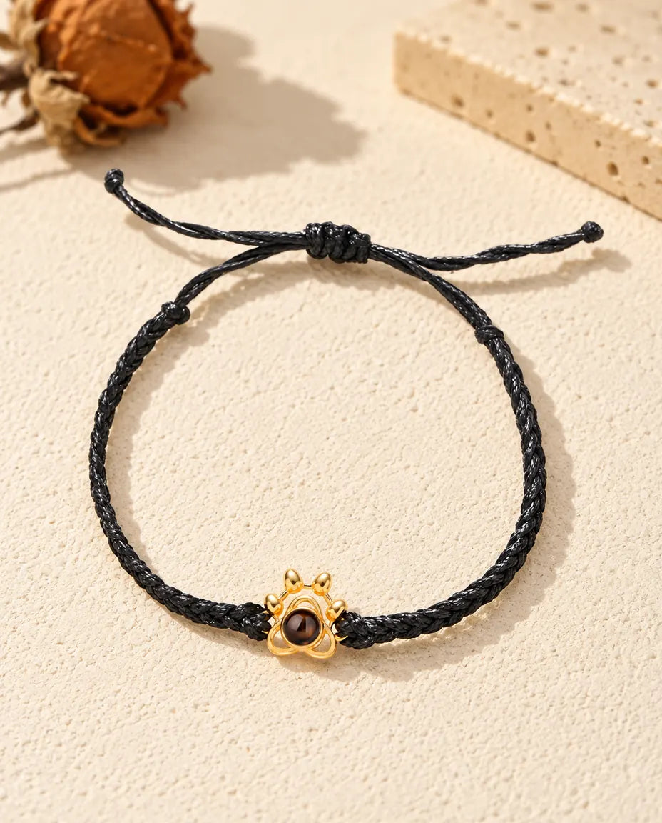 Bovenaanzicht van de armband met goudkleurig pootje aan een zwart gevlochten koord en de schuifknoop