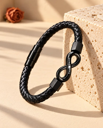 Zwarte armband van gevlochten kunstleer met gegraveerd oneindigheidsteken, rechtop tegen een blok travertijn