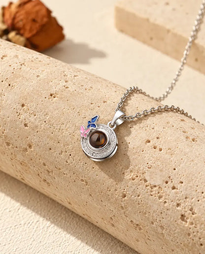 Gepersonaliseerde zilverkleurige ketting met rond hangertje en donkerblauwe en roze emaille vlindertjes, op travertijn