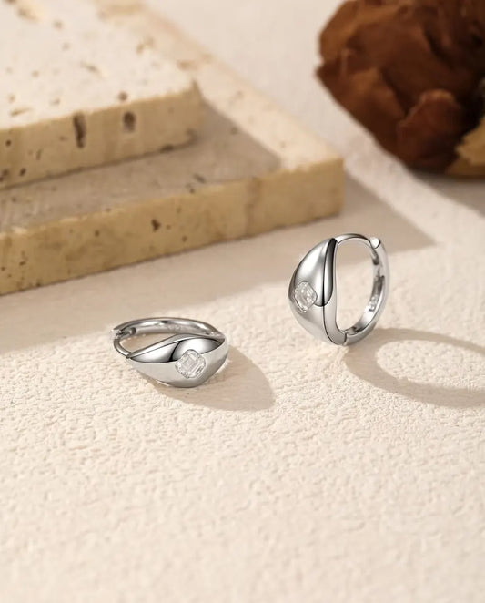 Moissanite Huggie Creolen met Asscher Slijpsel in 925 Sterling Zilver - het paar op een licht gestructureerd oppervlak, één liggend en één staand